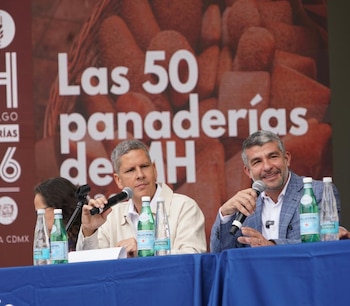 Dos hombres sentados en una mesa azul con micrófonos y botellas de agua frente a un cartel rojo con la frase "Las 50 panaderías de MH"