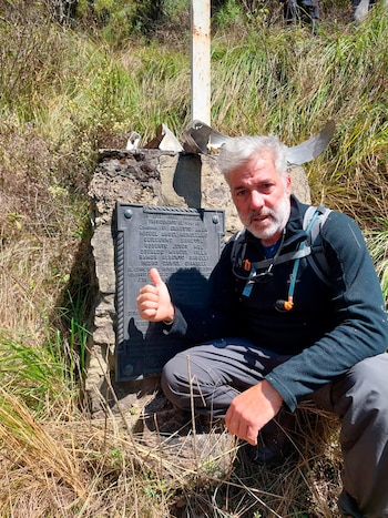 Hombre de barba y cabello gris, con ropa deportiva y mochila, agachado junto a un monumento de piedra con placa, dando el pulgar arriba