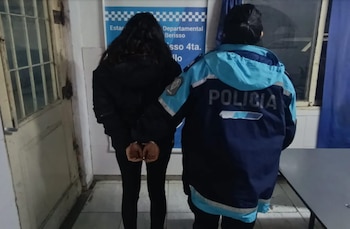 Mujer de espaldas con esposas en las manos, acompañada por un agente de policía con uniforme azul. Al fondo, una pared blanca con un cartel y una puerta de rejas