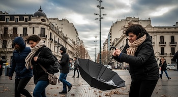 Una mujer con un paraguas negro invertido por el viento en una calle urbana. Otras personas abrigadas caminan y hay edificios y hojas secas en el suelo.
