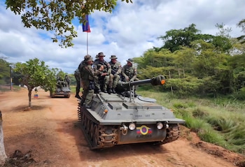 Cuatro soldados venezolanos uniformados sentados sobre un vehículo blindado con cañón en un camino de tierra, rodeado de árboles y con la bandera de Venezuela, bajo un cielo nublado