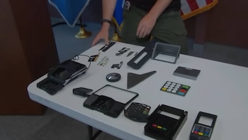 Una mano de persona, una mesa blanca, varios dispositivos electrónicos para clonación de tarjetas, piezas de terminales de pago, una lupa y una bandera