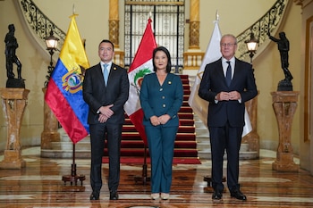 Tres personas de pie en un vestíbulo con escaleras alfombradas, junto a las banderas de Ecuador, Perú y la Comunidad Andina