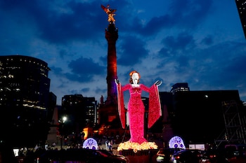 Una gran figura iluminada de color rosa con rostro de calavera se ubica frente a una columna coronada por una estatua dorada al anochecer
