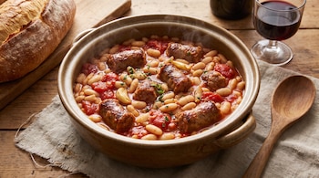Cazuela de barro con alubias blancas, tomate y longaniza, junto a un pan de hogaza, una copa de vino tinto y una cuchara de madera.