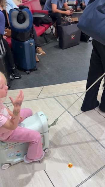 Una niña con ropa rosa va sentada sobre una maleta infantil con ruedas mientras una persona la remolca en la terminal de un aeropuerto