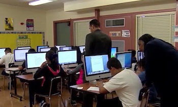 Un aula con mesas individuales y computadoras iMac, varios alumnos sentados frente a las pantallas, y dos adultos de pie observando