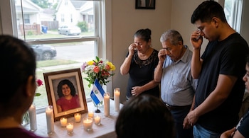 Tres personas adultas lloran junto a un altar con la fotografía enmarcada de una mujer sonriente, velas, flores y una bandera de El Salvador en una casa.