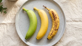 Tres plátanos de color verde, amarillo y marrón con manchas sobre un plato gris sobre una tela beige