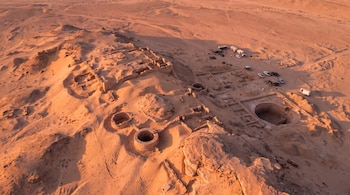 Vista aérea de paisaje desértico con ruinas, excavaciones arqueológicas, pozos circulares de piedra, estructuras enterradas, carpas y vehículos en un campamento.