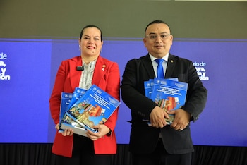 Una mujer con chaqueta roja y un hombre con traje oscuro sostienen libros azules impresos frente a una pantalla azul