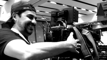 Primer plano en blanco y negro de Martín Guzmán sonriendo y mirando hacia la derecha, con una gorra, junto a una cámara profesional