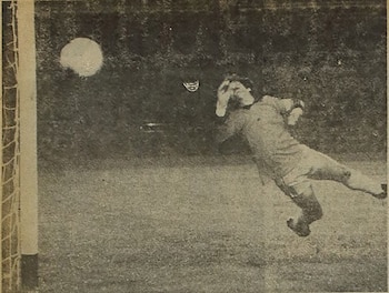 Imagen en blanco y negro de un portero de fútbol, vestido con uniforme claro, saltando con los brazos extendidos para atajar un balón cerca de un poste