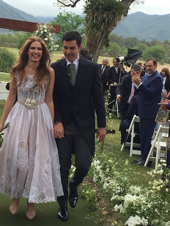 Un vestido midi en organza de seda natural off white, con importante corsage bordado con transparencias en hilos de seda con flores y rayas de vainillas. El detalle una hebilla en flores y hojas emulando el vestido, con los detalles de las iniciales de los novios realizado por el orfebre salteño Horacio Bertero