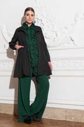 Impermeable entallado negro con bolsillos y detalle de cuello de encaje. Conjunto de blusa sin mangas con jabot y pantalón oxford de seda natural en verde esmeralda. En sus pies, loafers de raso sin talón. De accesorios, maxi aros doble círculo en metalizado y negro de Luna Garzón