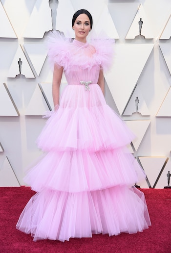 Kacey Musgraves eligió un diseño en color rosa bebé de tul en cascada con cinturón de aplique con brillantes en forma de moño by Giambastista Valli. Completó su beauty look con cabello recogido, smokey eye en negro y plateado y boca nude.