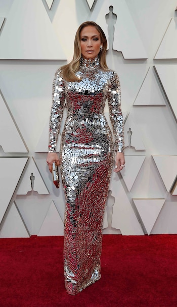 Jennifer Lopez enfundada en un vestido sirena con mangas largas y cuello polera de piedras plateadas. Completó el look con clutch rígido a tono y aros clip. Su beauty look, cabello lacio hacia un costado, smokey eyes y labios nude